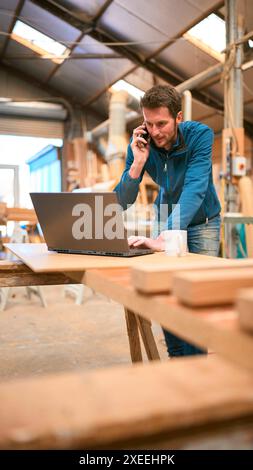 Carpenter Working In Woodwork Workshop Talking On Mobile Phone And Using Laptop Stock Photo