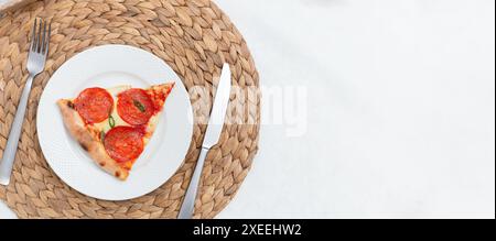 Pepperoni pizza slice banner on a white plate, placed on a woven placemat with a fork and knife Stock Photo