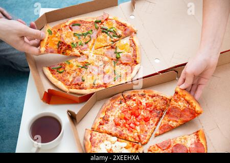 Two people grabbing slices of pizza from open delivery boxes with a cup of tea on a white table. Stock Photo