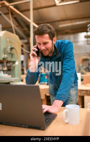 Carpenter Working In Woodwork Workshop Talking On Mobile Phone And Using Laptop Stock Photo