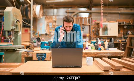 Carpenter Working In Woodwork Workshop Talking On Mobile Phone And Using Laptop Stock Photo