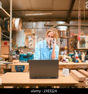 Female Carpenter Working In Woodwork Workshop Talking On Mobile Phone And Using Laptop Stock Photo