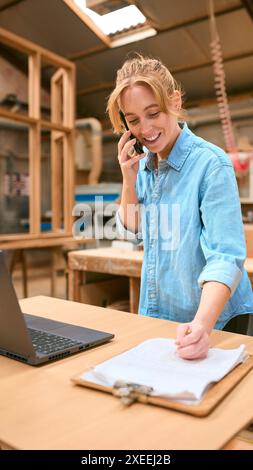 Female Carpenter Working In Woodwork Workshop Talking On Mobile Phone With Laptop Checking Order Stock Photo