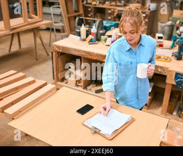 Female Carpenter Working In Woodwork Workshop With Cup Of Coffee Making Notes On Clipboard Stock Photo
