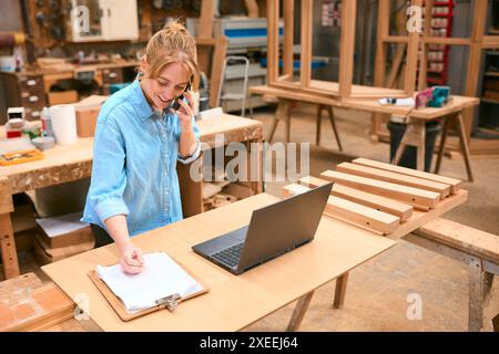 Female Carpenter Working In Woodwork Workshop Talking On Mobile Phone With Laptop Checking Order Stock Photo