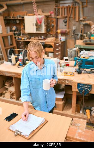 Female Carpenter Working In Woodwork Workshop With Cup Of Coffee Making Notes On Clipboard Stock Photo