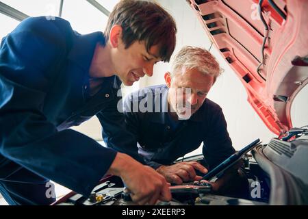 Male Car Auto Mechanic With Trainee Looking Under Bonnet Of Car Holding Digital Tablet Stock Photo