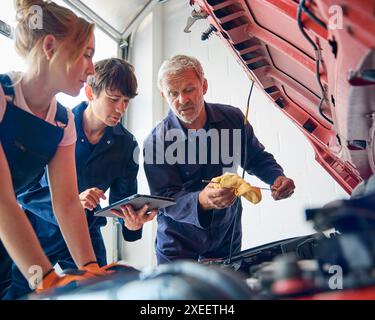 Team Of Car Auto Mechanics With Trainees Looking Under Bonnet Of Car Using Digital Tablet Stock Photo