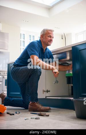 Mature Male Plumber Fixing Waste Disposal Unit In Domestic Kitchen Sink Stock Photo