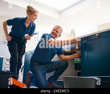 Male Plumber With Female Trainee Using Digital Tablet Fixing Waste Disposal Unit In Kitchen Sink Stock Photo