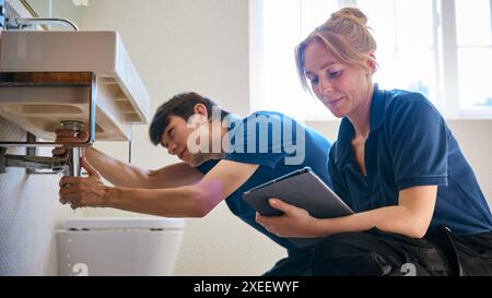 Female Plumber Holding Digital Tablet With Male Trainee Fixing Leak In Domestic Bathroom Sink Stock Photo