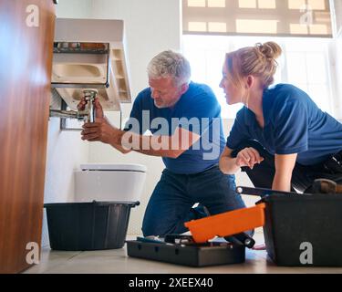 Male Plumber Fixing Leak In Domestic Bathroom Sink With Female Trainee Holding Digital Tablet Stock Photo