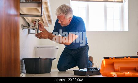 Mature Male Plumber Fixing Leak In Domestic Bathroom Sink Or Basin Stock Photo