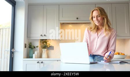 Smiling Woman Working From Home In Kitchen Using Laptop Stock Photo