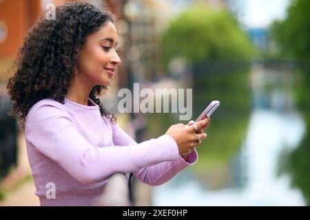 Young Woman Standing By River In Urban Area Using Mobile Phone For Messaging Or Social Media Stock Photo