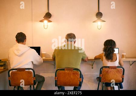 Rear View Of Three Businesspeople Sitting At Counter In Coffee Shop Working On Laptop Stock Photo