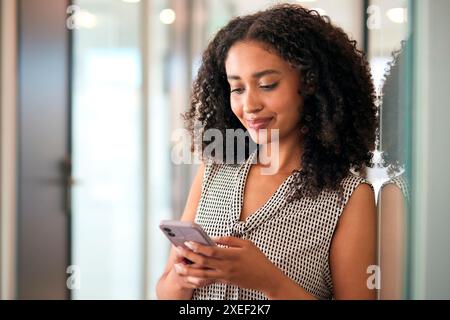 Businesswoman Standing In Corridor Of Modern Office Building Using Mobile Phone Stock Photo