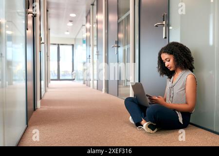 Businesswoman Sitting On Floor In Corridor Of Modern Office Working On Laptop Using Mobile Phone Stock Photo