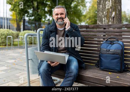 Portrait of a mature adult male director with a beard works using a laptop and speaks on the phone in the city Stock Photo