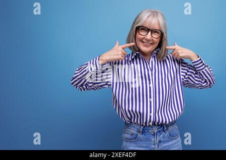 Smiling kind 60s female pensioner with gray hair in a stylish look with a grimace on a bright studio background with copy space Stock Photo