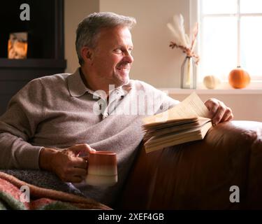 Mature Man At Home In Winter Jumper On Sofa With Warming Hot Drink Of Coffee In Mug Reading Book Stock Photo