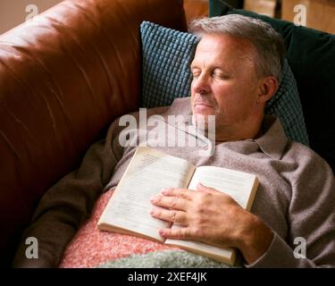 Mature Man At Home In Winter Jumper Sleeping On Sofa With Warm Blanket Holding Book Stock Photo