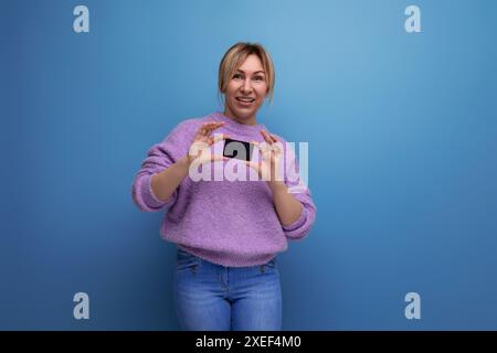 Adorable blonde young woman in a lilac sweater holding a plastic credit card mockup on a bright background with copy space Stock Photo