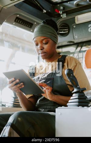 Vertical medium shot of young Black female factory worker sitting inside tractor using digital tablet Stock Photo