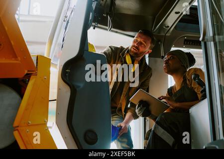 Low angle view shot of two ethnically diverse male and female factory workers checking quality of new bulldozer cabin Stock Photo