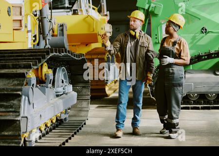 Long shot of two ethnically diverse male and female engineers walking along factory workshop and discussing new design of modern dozer Stock Photo