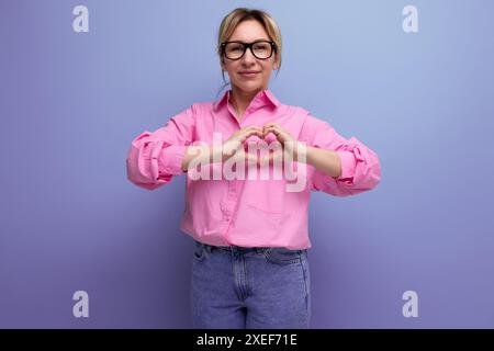 Young energetic caucasian blonde secretary woman with ponytail hairstyle, glasses and in a pink shirt shows a heart on a studio Stock Photo