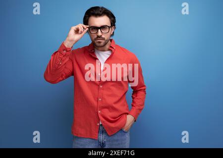 Surprised caucasian brunette 30s man in a red shirt holding glasses in urki on background with copy space Stock Photo