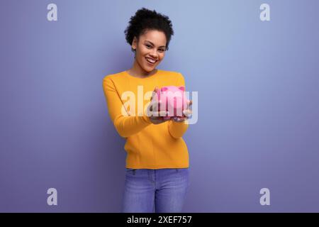Pretty joyful 20s latin woman with afro hair in casual yellow sweater accumulated money in a piggy bank Stock Photo