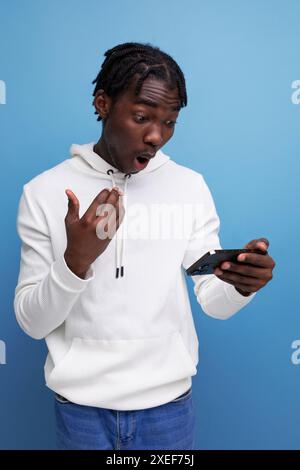 Surprised young african guy in a white sweatshirt understands the smartphone Stock Photo
