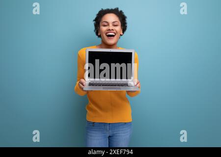 Pretty young brunette latin female adult in yellow sweater showing laptop screen with mockup for advertising Stock Photo