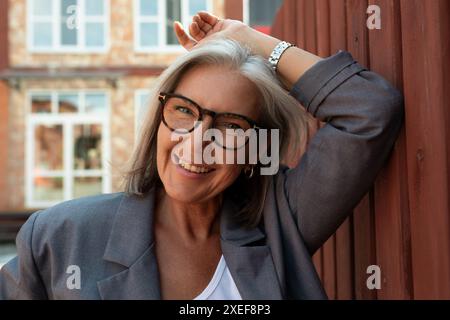 Stylish mature woman with gray hair and glasses is dressed in a gray jacket walking around the city Stock Photo
