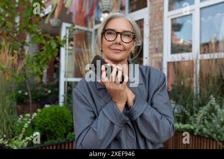 A well-groomed senior woman with gray hair and glasses is dressed in a gray jacket and jeans, holding a smartphone in her hand a Stock Photo