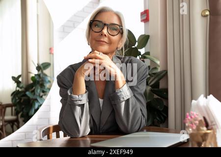 Confident well-groomed senior woman with gray hair and glasses dressed in a gray jacket sits in a cafe at a table with a laptop Stock Photo