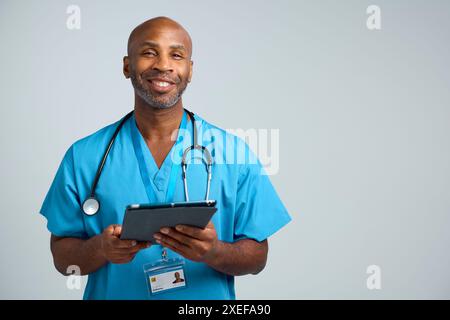 Studio Portrait Shot Of Smiling Mature Doctor Wearing Scrubs Using Digital Tablet Stock Photo