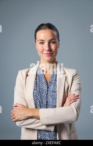 Studio Portrait Of Smiling Businesswoman Wearing Suit With Folded Arms Against Grey Background Stock Photo
