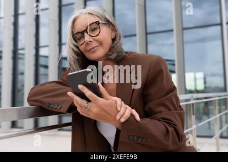 60 year old successful slender gray-haired business woman dressed stylishly checks messages on the mail on the phone Stock Photo