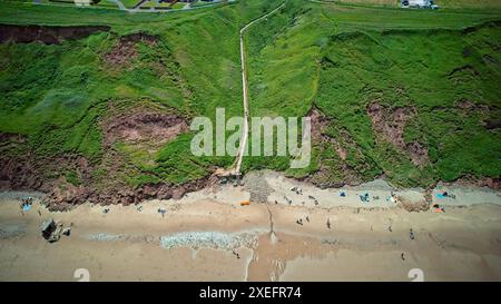 Aerial view of a coastal cliff with a staircase leading down to a sandy beach. The cliff is covered in green vegetation, and there are people on the b Stock Photo