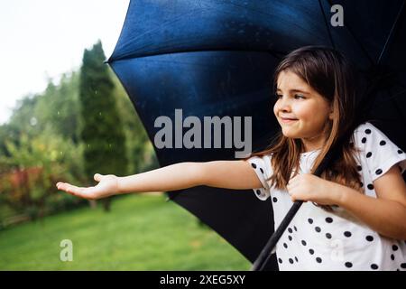 A little girl stands with a black umbrella in the rain. A cute baby stretches out her hand and catches raindrops in the park. Sweet happy smiling baby Stock Photo