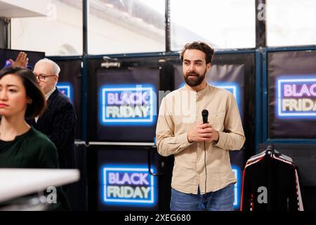 Young man local TV reporter holding mic doing live broadcast from fashion outlet during Black Friday sales. Male correspondent covering scene at shopping mall on busiest retail day of year Stock Photo