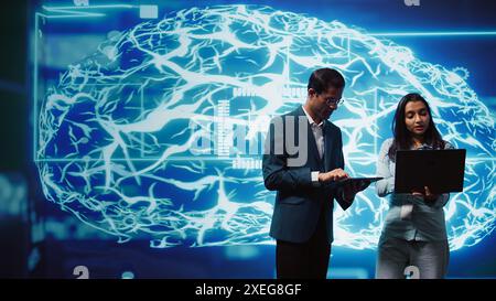 IT department colleagues solving tasks on laptop and tablet, running artificial intelligence programming scripts. Team of engineers brainstorming ways to develop AI applications, camera B Stock Photo