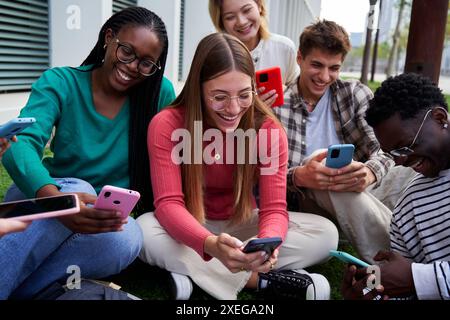 Group of friends gathering outdoors sitting on the ground using phones watching social media content Stock Photo