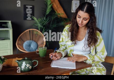 Charming curly-haired brunette in casual clothes signs documents at home. Young attractive woman fills out paper forms in living Stock Photo