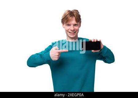 Young handsome red-haired man with a blue sweater points a finger at a smartphone Stock Photo