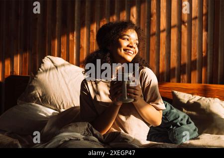 Charming mixed-race teenager is laughing and holding cup of coffee while sitting in bed. Pretty African girl is enjoying sunny m Stock Photo