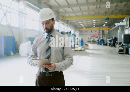 Bearded Caucasian engineer in formalwear with hardhat standing in factory workshop working on digital tablet, copy space Stock Photo